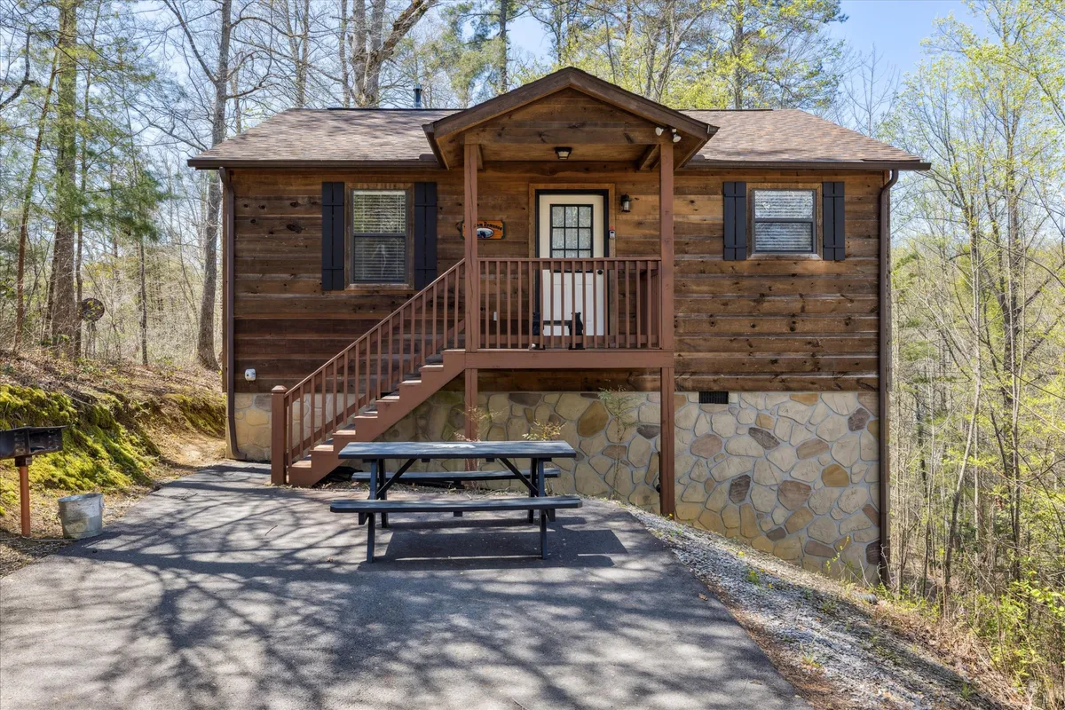 The image features the charming wooden exterior of a Smoky Mountains cabin, set atop a sturdy stone foundation. A welcoming staircase leads up to a spacious porch, while a picnic table invites guests to dine outdoors amid the peaceful, wooded surroundings. The mood is relaxed, perfect for enjoying sunsets, stargazing, or morning coffee on the porch.