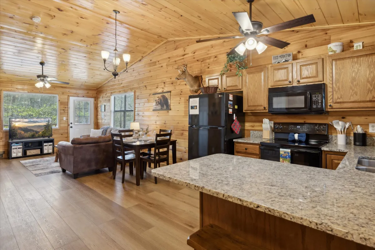 An inviting, open-concept space with a granite kitchen countertop, wooden dining table, refrigerator, and a view into the living area with a comfortable couch. Warm wood finishes and rustic touches tie the kitchen, dining, and living areas together seamlessly.