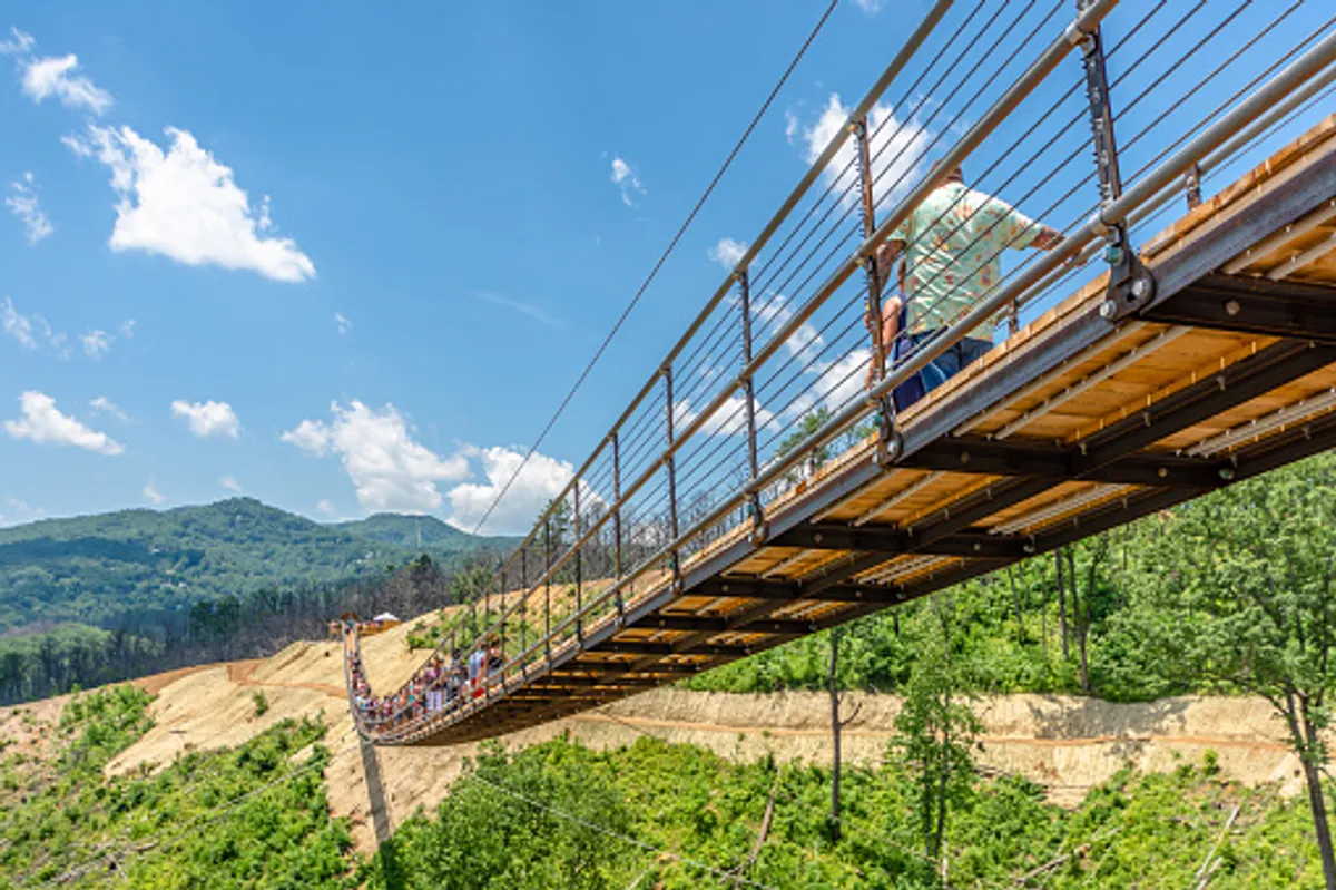 A thrilling suspension bridge stretches across a lush green landscape, with people walking across and majestic mountains providing an awe-inspiring backdrop. The sense of adventure is palpable, signaling opportunities for couples to explore, hike, and connect with nature.