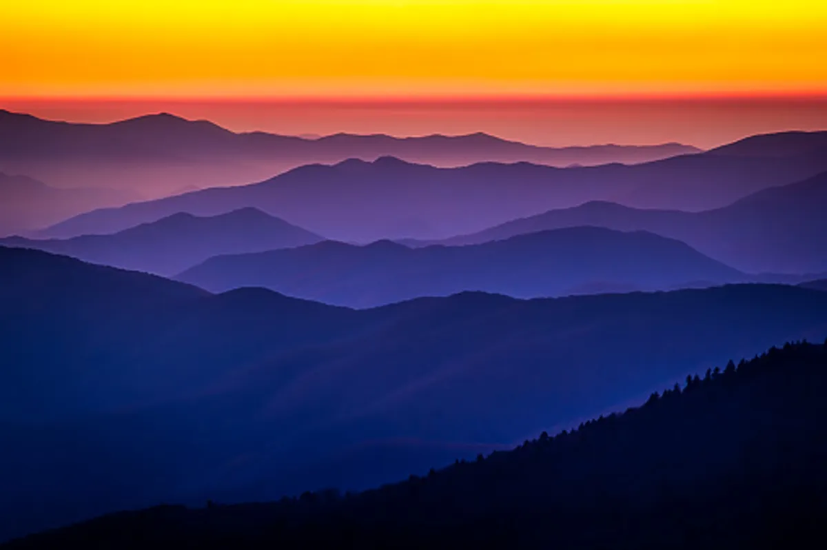 This image showcases a breathtaking view of layered mountain ranges bathed in the vibrant hues of a sunset. The sky is awash with warm colors, and the serene landscape invites reflection and relaxation, perfectly evoking the tranquil beauty of the Smoky Mountains at dusk.