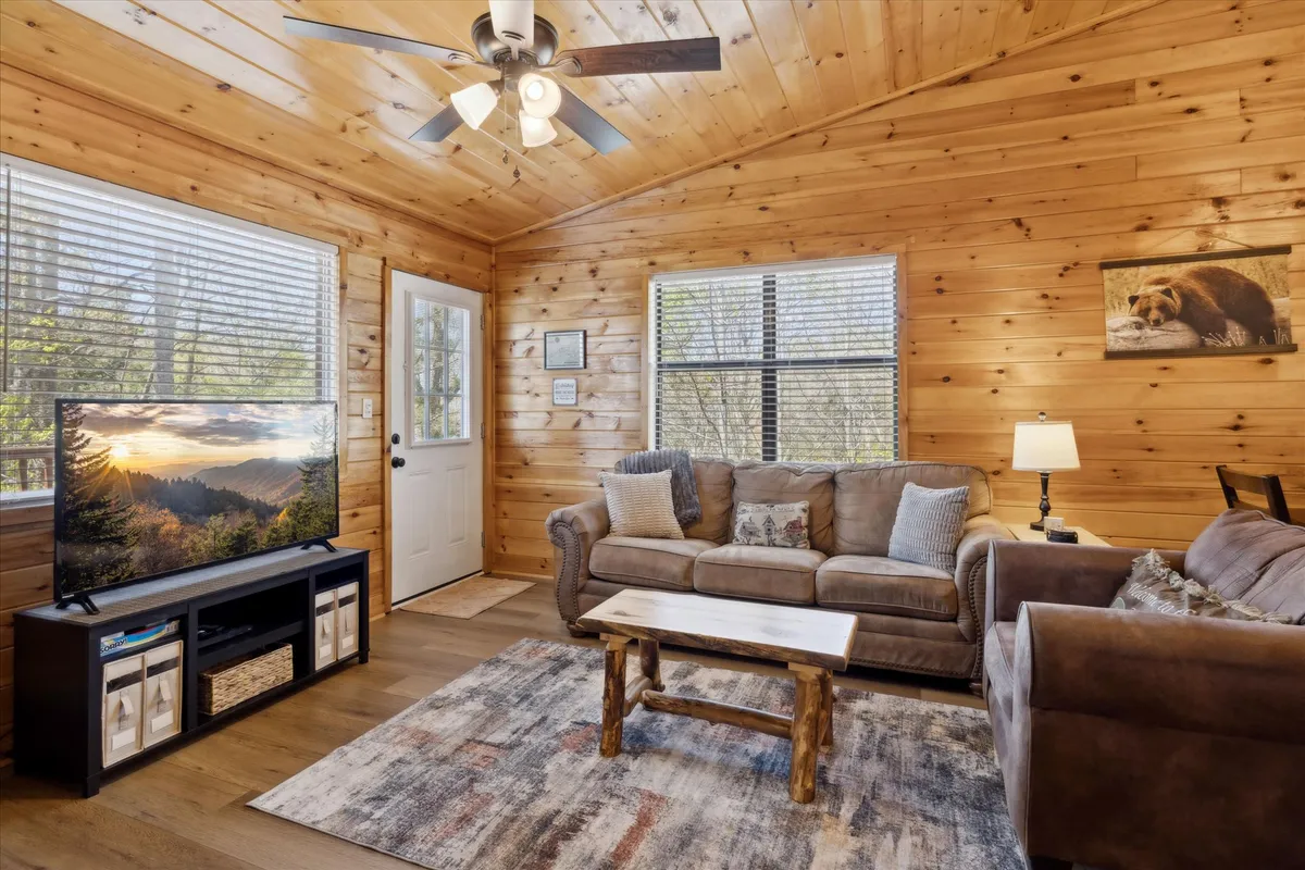 This warm, inviting living room features a comfortable sofa, a wooden coffee table, and a large TV displaying a scenic view. The natural wooden interiors and the soft light streaming through the windows create an atmosphere of relaxation, suggesting a cozy retreat after a day of adventure.