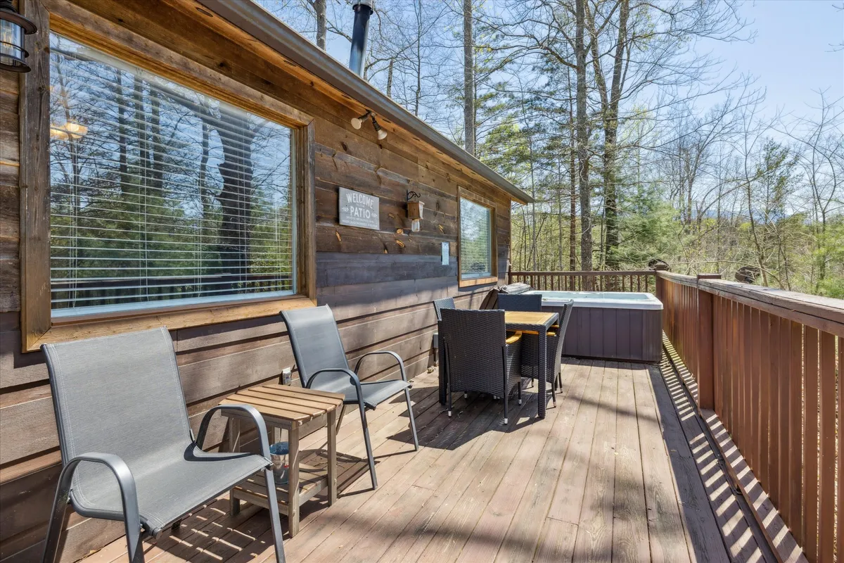 A spacious wooden deck with a hot tub and comfortable outdoor seating set against the backdrop of a lush forest. The deck invites al fresco dining or quiet mornings with coffee, while the hot tub promises warm relaxation under the sky. The cabin's wooden siding and generous windows add to the inviting, nature-connected vibe.