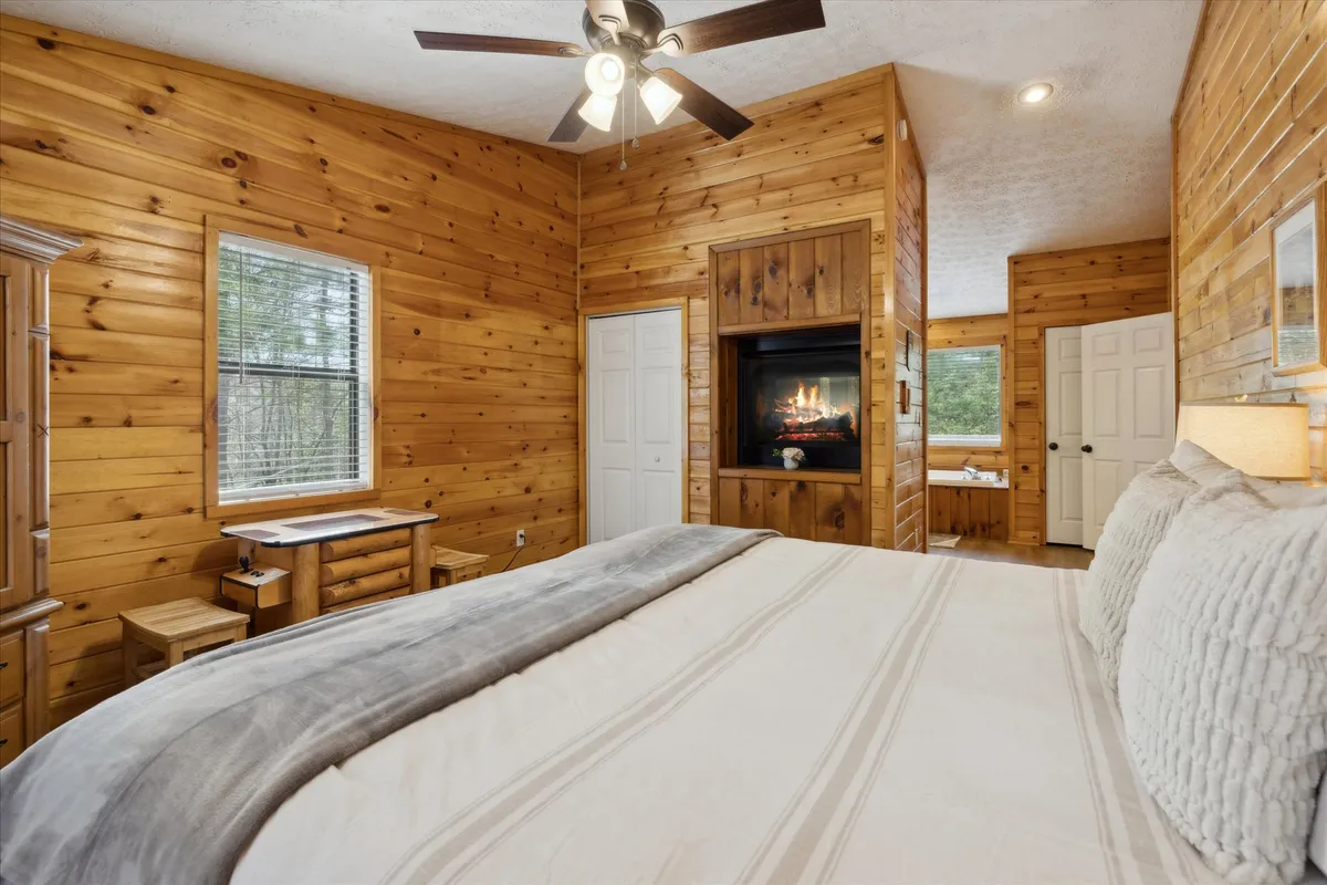 A beautifully appointed bedroom featuring a plush king bed, elegant wood paneling, and a cozy fireplace. Soft, ambient light filters in through the window, casting a romantic glow across the room and emphasizing the warmth of the fireplace.