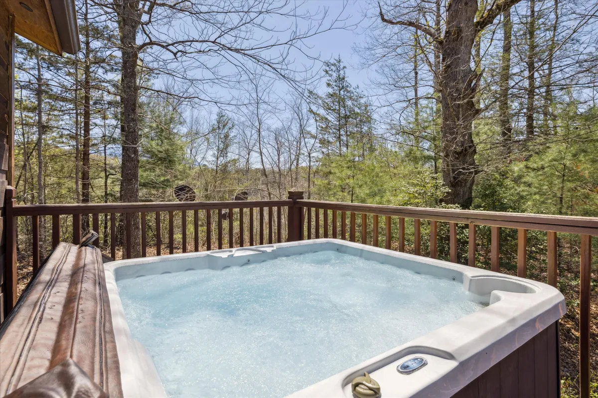 This image showcases a serene outdoor deck, featuring a bubbling hot tub nestled among towering trees. The deck railing frames the view of the woodland, and the hot tub is positioned to maximize privacy and immersion in nature. The setting exudes tranquility, perfect for relaxation and enjoying the fresh mountain air.