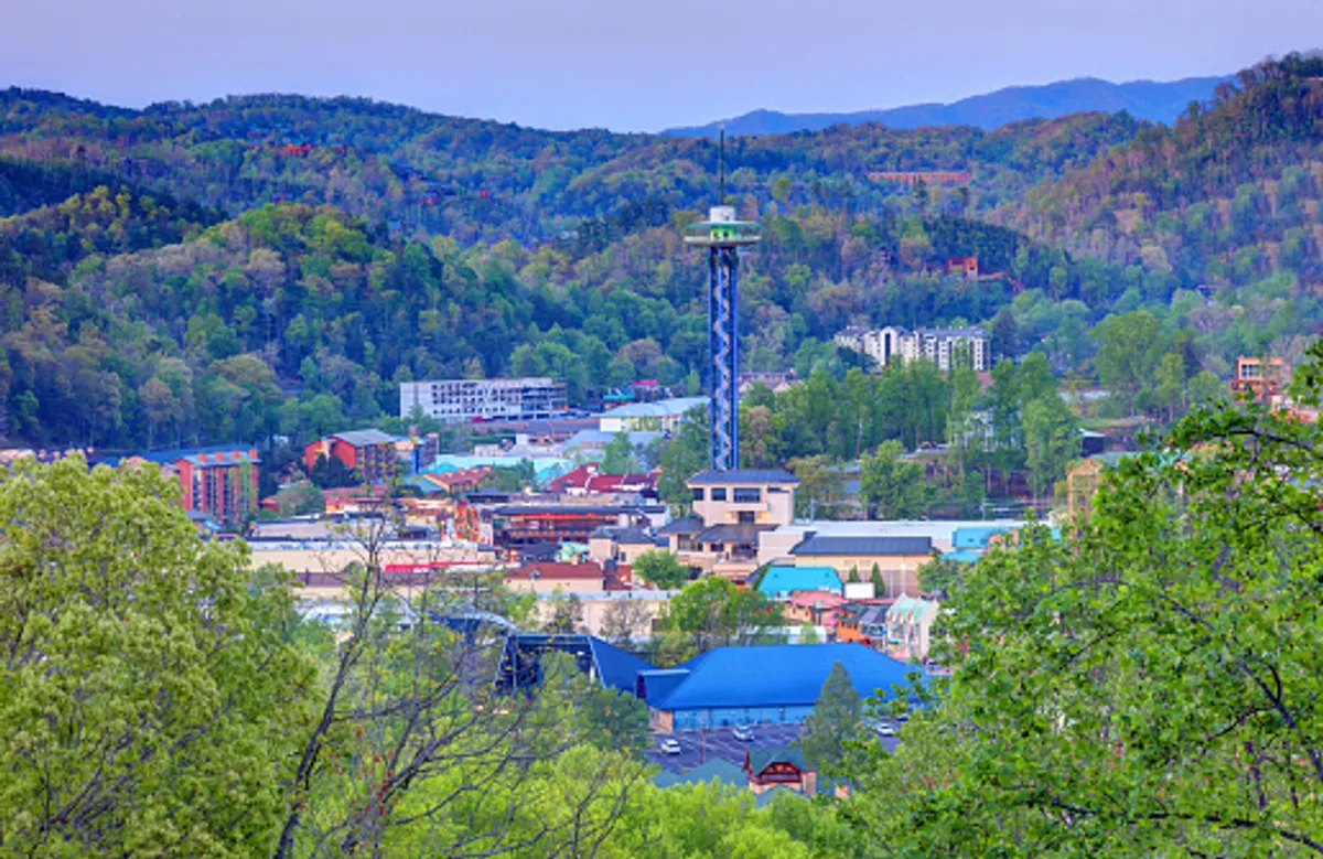 A panoramic daytime view of the town nestled below a majestic mountain range, featuring an observation tower and colorful buildings. The scene showcases the vibrant energy of the area and suggests a range of activities, from sightseeing to exploring local shops and eateries.
