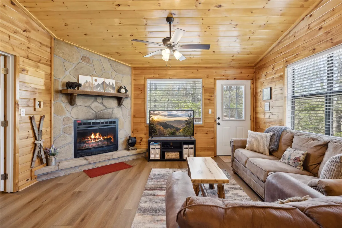 A warmly lit living area featuring a plush sofa, a glowing double-sided fireplace, and a television. Large windows let natural light pour in, framing the surrounding woodland scenery. The wood finishes and soft furnishings create a cozy, intimate ambiance ideal for couples.